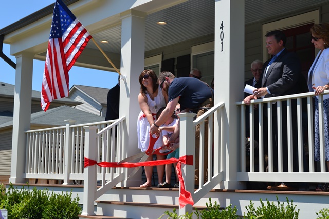 Operation Finally Home Ribbon Cutting Family cutting a giant red ribbon on a home porch with an American Flag