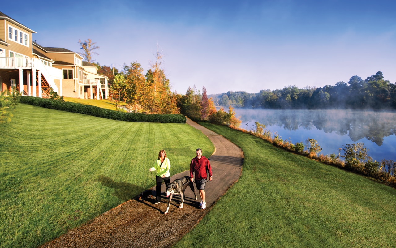 A couple walking a dog along a dirt path in the morning with homes on a small hill on the left and a lake on the right