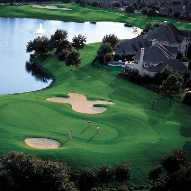 Aerial of three people on the green of a golf course with a pond in the background