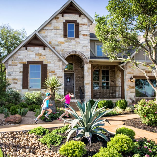 Two kids running on a sidewalk in front of a stone home with shrubs all around